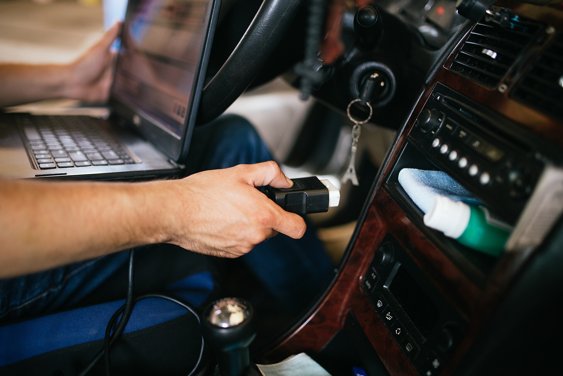 Een hand steekt de uitleesstekker van een diagnoseapparaat in een auto, naast een laptop
